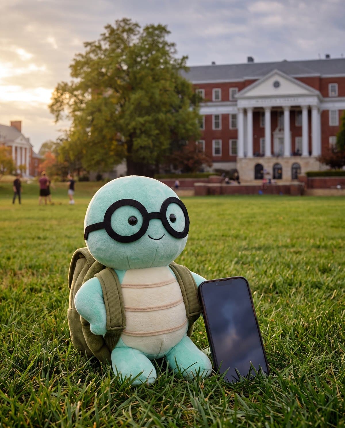 The NoTestudo plush on the grass of McKeldin Mall beside a phone, a brick building with white columns behind it.